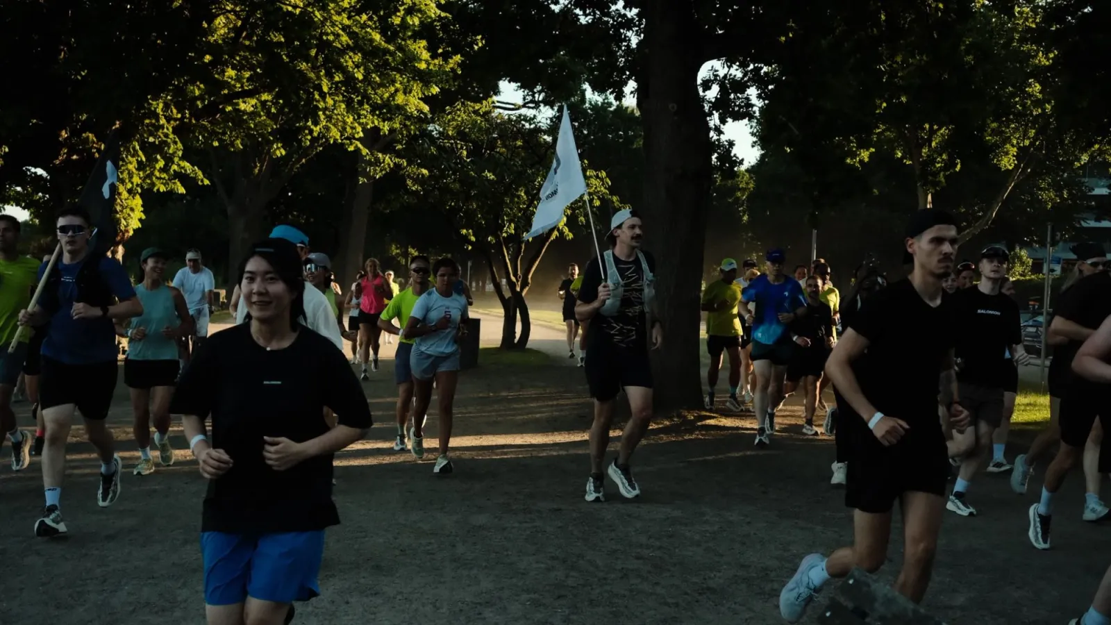 Läuferinnen und Läufer eines Community-Runs im Park, im Abendlicht, vorne eine lachende Läuferin, dahinter ein Läufer mit weißer Fahne